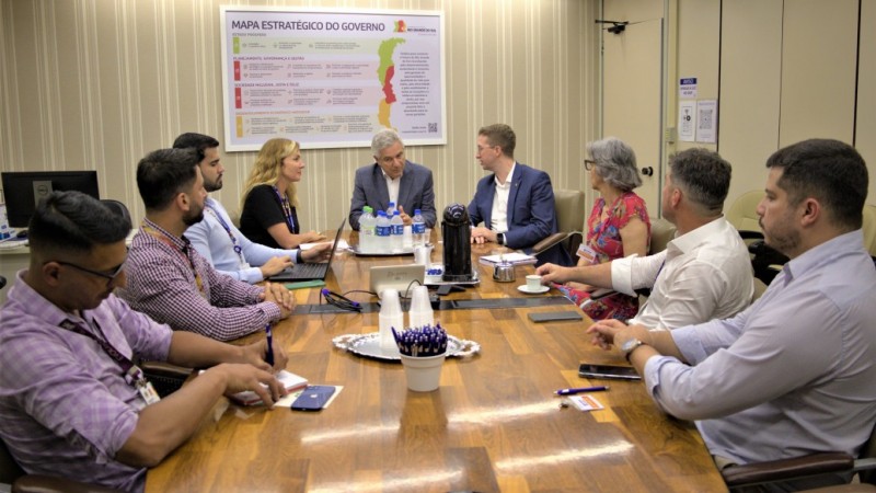 Sala de paredes predominantemente amarelas, com pessoas sentadas ao entorno de uma mesa de reunião.